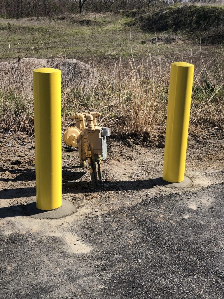 Yellow bollards installed around utility equipment on a gravel and asphalt surface.